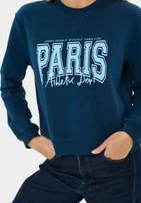 Felpa blu navy con il testo "PARIS Athletic Dept.", indossata con le mani nelle tasche di jeans scuri.
