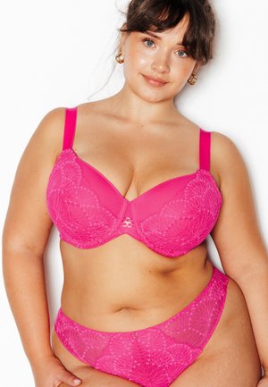 Femme portant un soutien-gorge en dentelle rose vif assorti et une culotte taille haute, avec des boucles d'oreilles en or en forme d’anneau sur fond blanc.