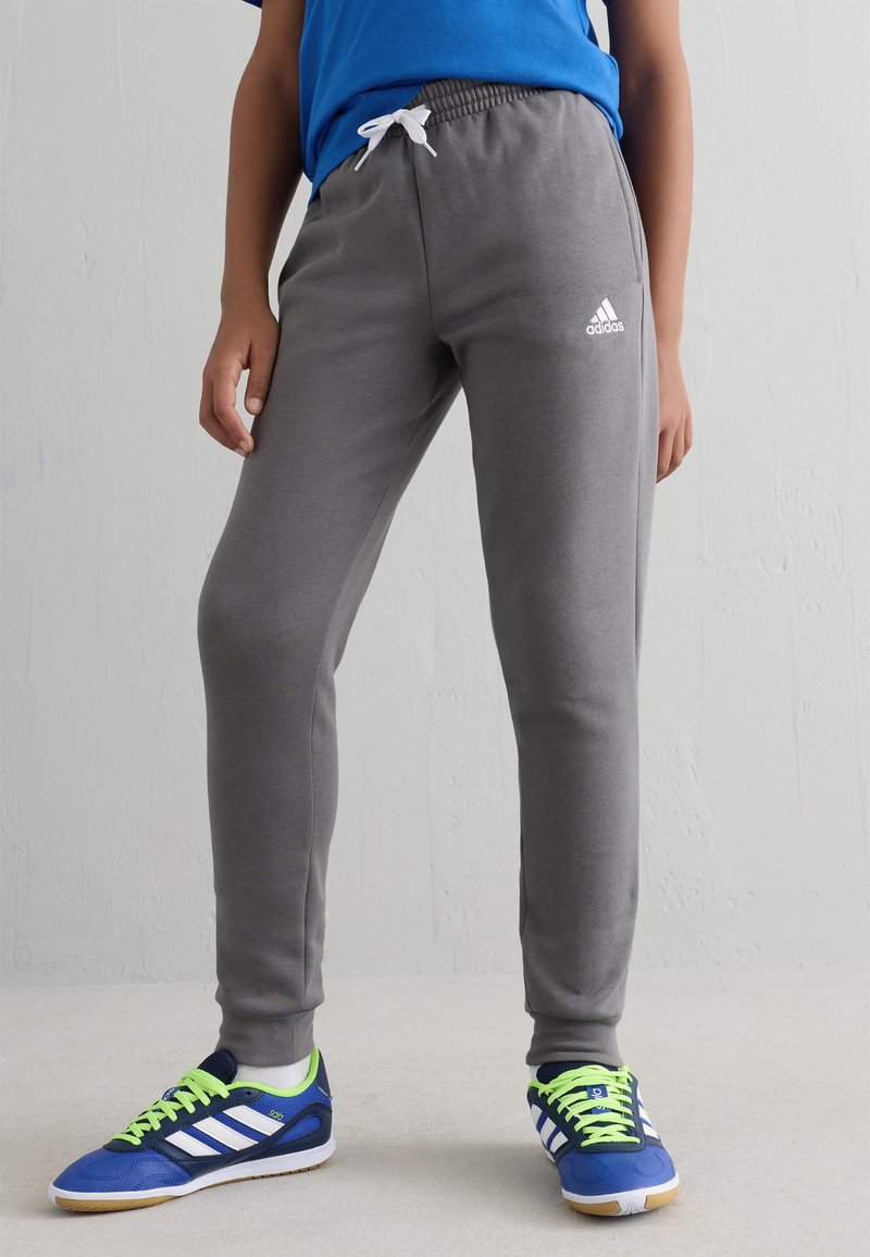 Sive trenerke s elastikim pojasom i ušicom. Na njima se nalazi Adidas logo u bijeloj boji. Uparene su s plavim sportskim cipelama i neonsko zelenim pertlama.