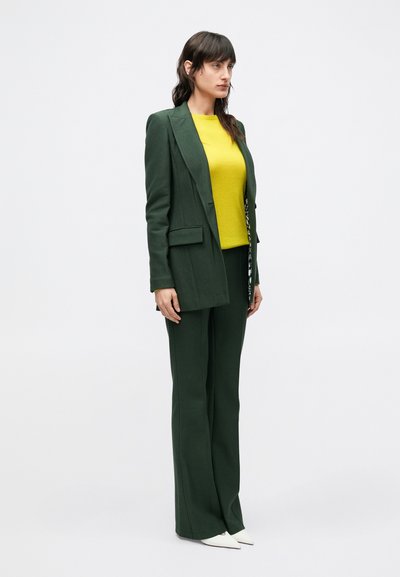 Zelený mierne priliehavý oblek s štruktúrovaným sako a nohavicami so širokými nohavicami, doplnený jasne žltým topom a bielymi topánkami so špicatou špičkou.
