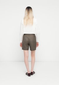 Vrouw met lang blond haar, gekleed in een witte blouse met lange mouwen, bruine high-waist shorts en donkerbruine platte schoenen, staand tegen een witte achtergrond.
