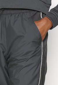 Černé lehké sportovní kalhoty s hladkou texturou, elastickým pasem, boční kapsou a kontrastním bílým lemováním podél švů.