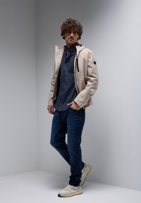 Chaqueta ligera beige con cierre en la parte delantera, acompañada de una camisa azul oscura de botones y jeans azules. Los zapatos son claros con acentos oscuros.