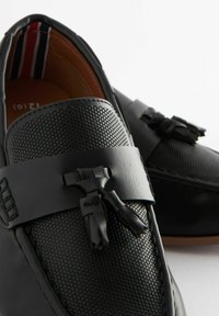 Mocassins noirs avec une tige en cuir perforé texturé, une sangle en cuir lisse et des accents de glands ; doublure intérieure à rayures discrètes.