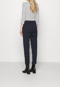 Vrouw met lang blond haar staat met haar rug naar ons gekeerd, gekleed in een wit en zwart gestreept shirt, marineblauwe broek en zwarte enkellaarsjes.