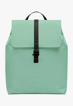 Mäta zelený batoh so zapínaním na klopu, čiernym popruhom a prackou, hladkou textúrou a minimalistickým dizajnom. Logo značky tlačené na spodnej strane.