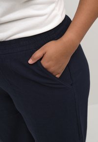 Námořnické modré zúžené kalhoty s texturovaným povrchem, s elastickým pasem a bočními kapsami. Ruka odpočívá v levé kapse.
