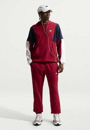 Mand iført en vinrød Nike træningsdragt med marineblå og hvide ærmer, hvid Nike kasket, hvide sneakers og høretelefoner, står på en ensartet lys baggrund.