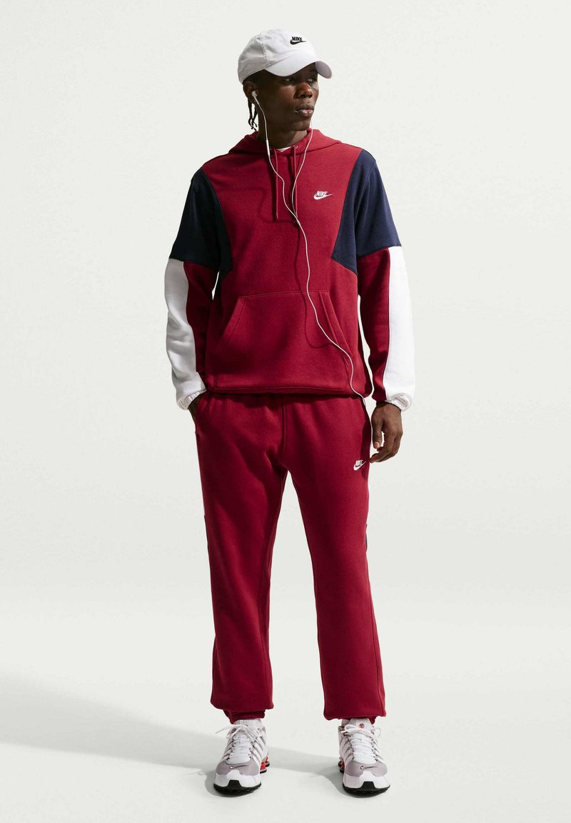 Uomo con tuta Nike color bordeaux con maniche blu navy e bianche, cappellino Nike bianco, scarpe da ginnastica bianche e auricolari, in piedi su sfondo chiaro uniforme.