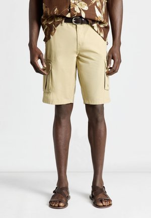 Uomo che indossa pantaloncini cargo beige con tasche laterali, una camicia marrone a fiori infilata e sandali di pelle marrone, in piedi su un pavimento bianco.
