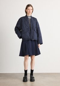 Marinblå quiltad jacka med rund halsringning, i kombination med en marinblå herringbone kjol. Modellen bär svarta platåskor och svarta strumpor.