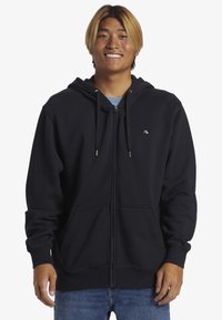 Sort zip-up hoodie lavet af blødt materiale, med en kængurulomme, hætte med snor og logo på brystet. Båret med blå jeans.