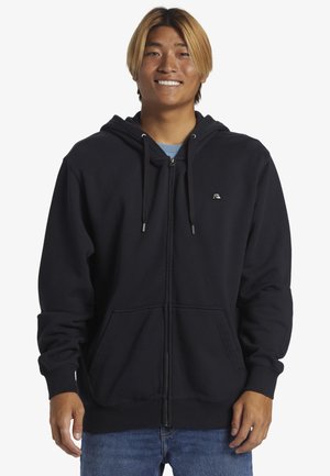 Sort zip-up hoodie lavet af blødt materiale, med en kængurulomme, hætte med snor og logo på brystet. Båret med blå jeans.