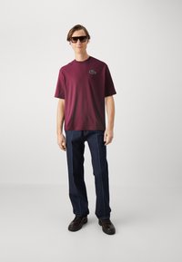 T-shirt di cotone bordeaux con maniche corte e logo ricamato verde, abbinata a jeans di denim scuro con cuciture chiare.