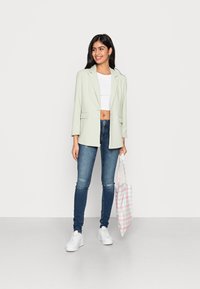 Blazer verde claro com lapelas em bico, combinado com uma t-shirt cropped branca e jeans skinny azuis. Leva uma bolsa de palha xadrez em tons pastel. Ténis brancos.
