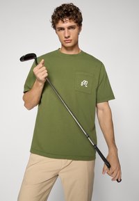 Olive green cotton T-shirt with a left chest pocket and white logo detail. Model holding a black golf club, wearing light beige pants.