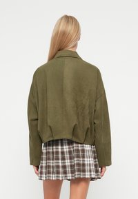Giacca in suede verde con una vestibilità loose e un design leggermente cropped, abbinata a una gonna a pieghe a scacchi con motivi marroni e bianchi.