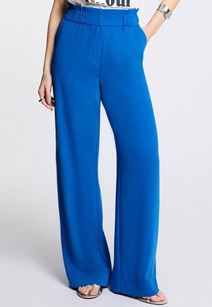 Blue wide-leg pants with elastic waistband worn by person standing with hands in pockets, paired with silver sandals and a cuff bracelet.