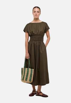 Femme debout portant une robe midi olive foncée avec taille froncée, tenant un sac tissé beige et vert à rayures et portant des sandales marron foncé.