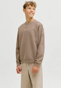 Teismeline poiss seisab ja naeratab, seljas taupe värvi dressipluus, millel on kiri "JACK & JONES" ning heledat beeži värvi püksid, tavalise tausta ees.