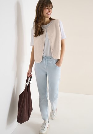 Kobieta uśmiechająca się, ubrana w beżową kamizelkę bez rękawów na białej koszulce i jasnoniebieskie spodnie ze sznurkiem, niosąca dużą ciemnobrązową torbę typu tote.