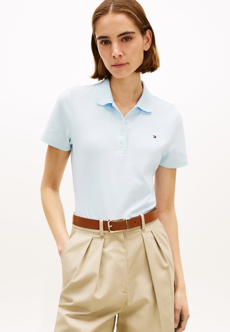 Femme portant un polo bleu clair rentré dans un pantalon plissé beige taille haute avec une ceinture marron, debout sur un fond blanc.