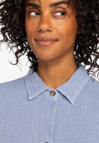 Light blue, textured fabric shirt with a buttoned collar, featuring a slightly wrinkled pattern and round button detail at the neck.