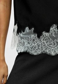 Haut en tissu noir avec un bord en dentelle blanche festonnée ornée de motifs floraux et de détails complexes le long de l'ourlet. Matière texturée visible.