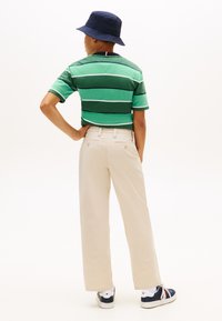 Άτομο που φοράει πουκάμισο με πράσινες ρίγες, μπεζ παντελόνι, μπλε καπέλο τύπου κάπα και αθλητικά παπούτσια, στέκεται με ένα χέρι στη μέση, στραμμένο προς τα πίσω.