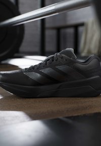 Μαύρο αθλητικό παπούτσι με τρεις ρίγες σε πρώτο πλάνο πάνω σε δάπεδο γυμναστηρίου κοντά σε μπάρα με βάρη άρσης βαρών.