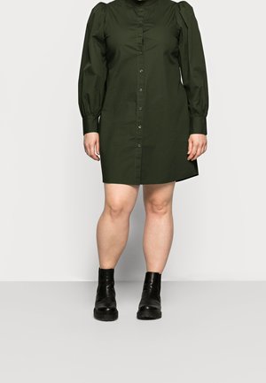 Femme portant une robe boutonnée olive à manches longues avec des épaules bouffantes et des bottines noires, sur un fond uni.