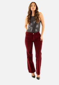 Pantaloni a zampa d'elefante in velluto rosso con tasche frontali, abbinati a un top senza maniche grigio lucido. La modella indossa tacchi a punta neri.