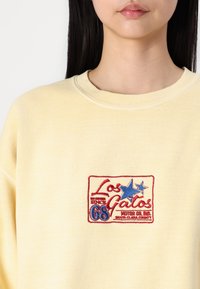 Gul sweatshirt med rund halsringning, med en färgglad broderad logotypdesign som inkluderar stjärnor och text på framsidan.