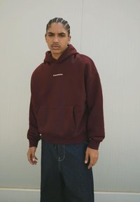 Giovane con capelli intrecciati che indossa un hoodie bordeaux con il testo "OVRNRM", jeans larghi neri, in piedi contro un muro grigio chiaro.