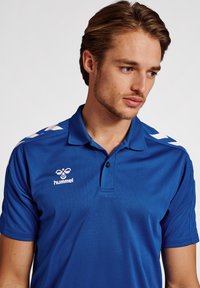Blå polo t-shirt med tekstureret stof, med knaplukning, hvide detaljer på skuldrene og et logo på venstre bryst.