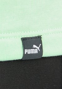 Liten svart tygetikett med vit Puma-logotyp och text fäst vid ljusgrön ribbad tyg ovanför svart texturerat tyg.
