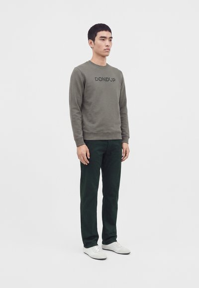 Homme debout portant un sweat-shirt vert olive avec le texte "DONDUP", pantalon vert foncé et baskets blanches, faisant face légèrement à droite.