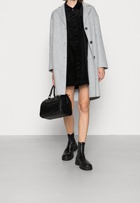 Cappotto di lana grigio, vestito in velluto a coste nero, borsa nera con motivo trapuntato e stivaletti neri chunky alla caviglia. Design minimalista, colori neutri.