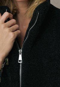 Zwarte fleecejas met een hoge kraag en zilverkleurige ritssluitingen. Zachte textuur en een nauwsluitende pasvorm zichtbaar rond de hals en het ritsgebied.