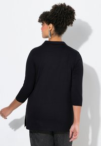 Tunica nera con colletto, maniche a 3/4 e spacchi laterali. Realizzata in un tessuto liscio e morbido. La modella indossa orecchini metallici.