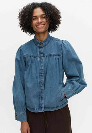 Smilende kvinde med krøllet hår iført en knappet blå denimjakke og mørke bukser med den ene hånd i lommen.