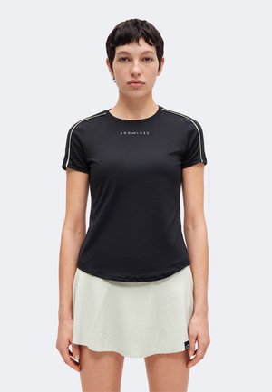 Mujer joven con cabello corto y oscuro, vistiendo una camiseta negra "ENDLESS" y una falda clara con textura, de pie frente a un fondo sencillo.