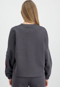 Sweatshirt gris à col ras du cou avec des manches longues et des poignets élastiques. Il dispose d'une poche sur le bras gauche et d'une étiquette cousue à l'arrière.
