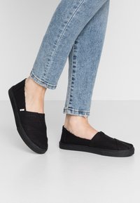 Svarta textilskor utan snörning med en strukturerad ovandel och en platt gummisula, bärs med uppvikt ljusblå jeans, vinklad mot betraktaren.