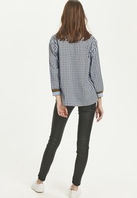 Kvinna med rakt brunt hår iförd svarta läderleggings, vita sneakers och en blåmönstrad långärmad topp, stående vänd bort. 