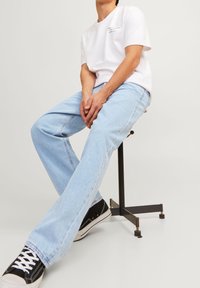 Vit bomullströja med ett litet motiv, ljusblå raka jeans och svarta sneakers med vita snörningar på en metalluppsats.