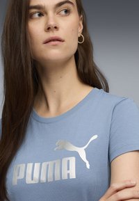 Šviesiai mėlynas trumpomis rankovėmis marškinėliai su dideliu sidabriniu "PUMA" logotipu ir puma grafika, pagaminti iš švelnios medvilnės medžiagos.