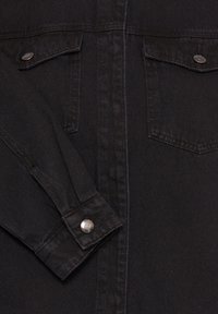 Veste en denim noire à l'avant avec deux poches poitrine boutonnées et un poignet boutonné sur la manche gauche posée sur le corps.