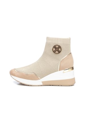 Beige strikket ankelstøvle med kilehæl, ruskindspynt på tå og hæl, elastisk manchet og dekorativt rundt metalemblem på siden.