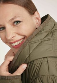 Kurtka pikowana w oliwkowym kolorze z kołnierzem stojącym, o gładkiej teksturze i detalach na zamek. Model trzyma kołnierz, uśmiechając się.